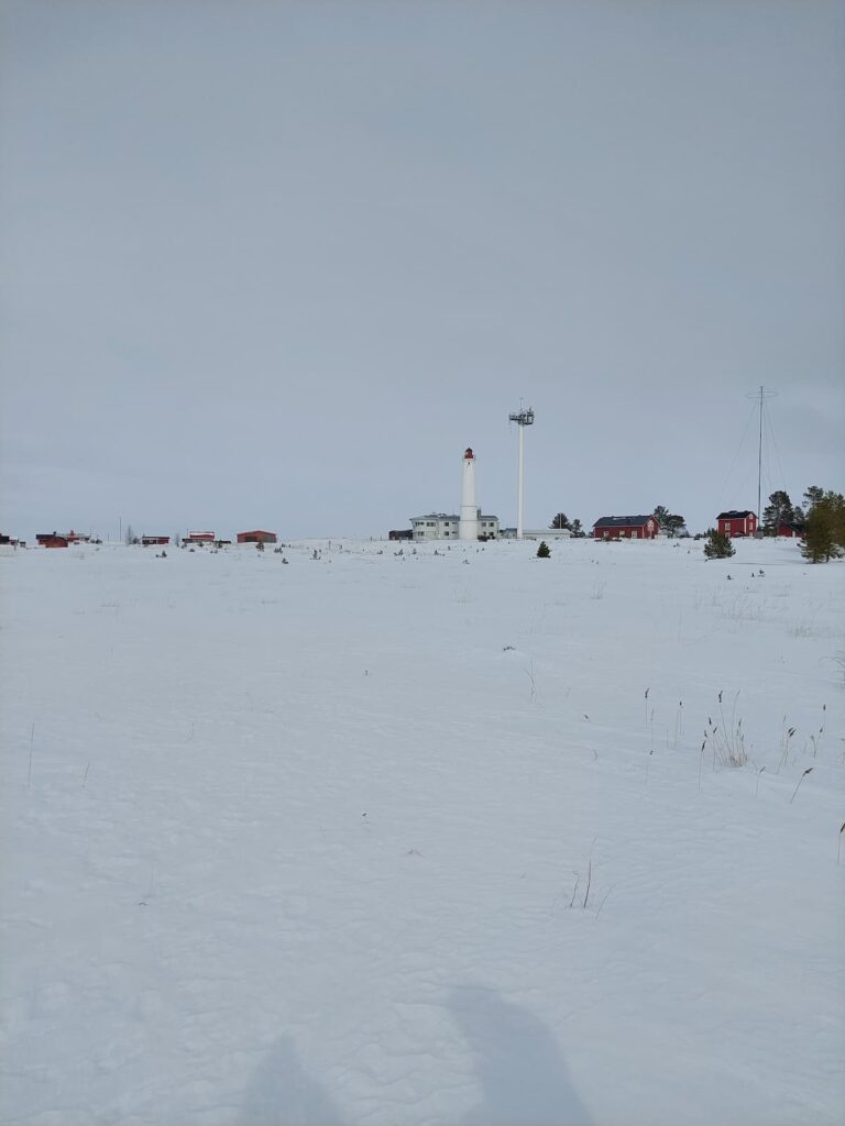 En vit fyr och flera röda byggnader står i ett snötäckt landskap under en molnig himmel, med glesa grässtrån i förgrunden.