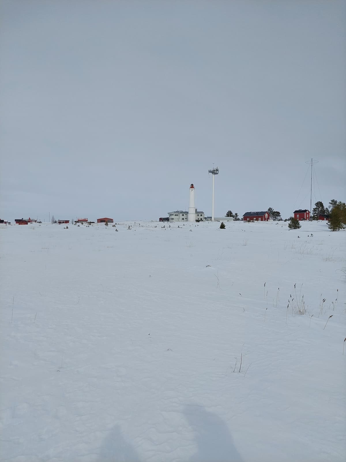 En vit fyr och flera röda byggnader står i ett snötäckt landskap under en molnig himmel, med glesa grässtrån i förgrunden.