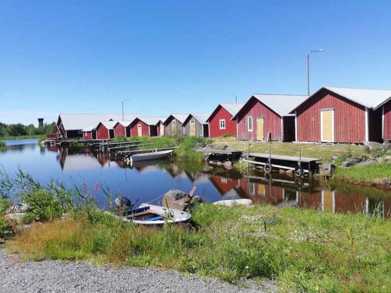 Rad av röda trästugor vid lugnt vatten med små båtar förtöjda längs stranden, grönt gräs i förgrunden och klarblå himmel ovanför.