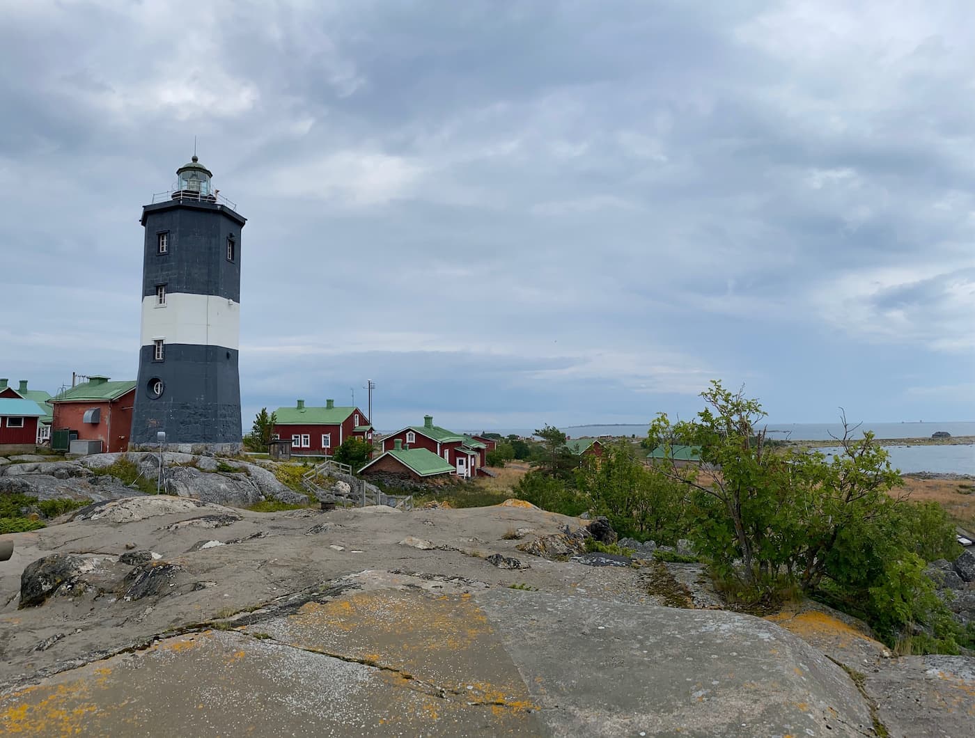 En svartvit fyr står på en klippig plats nära flera röda byggnader med gröna tak, med utsikt över havet under en molnig himmel.