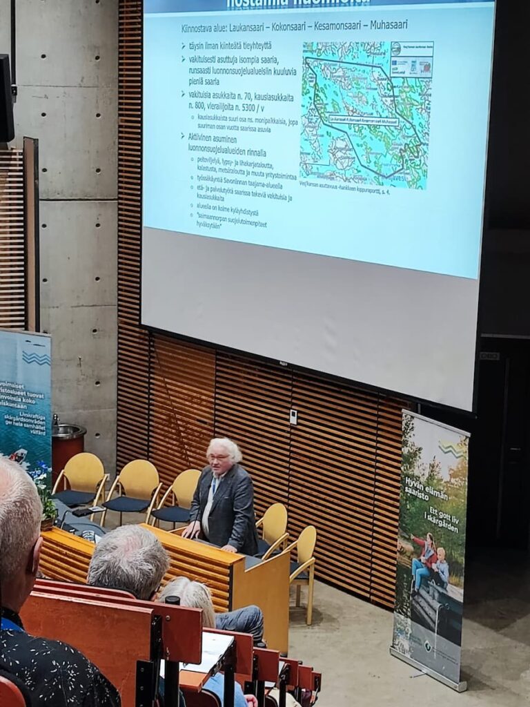 En man står på ett podium och håller en presentation i en föreläsningssal, med en projicerad bild som visar kartor och text på finska. Åhörarna sitter ner och tittar på.