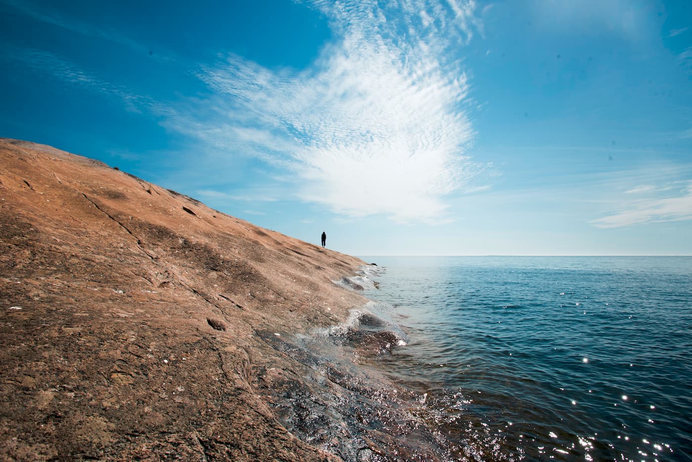 En person står ensam på en stenig strand vid det lugna blå vattnet under en delvis molnig himmel.