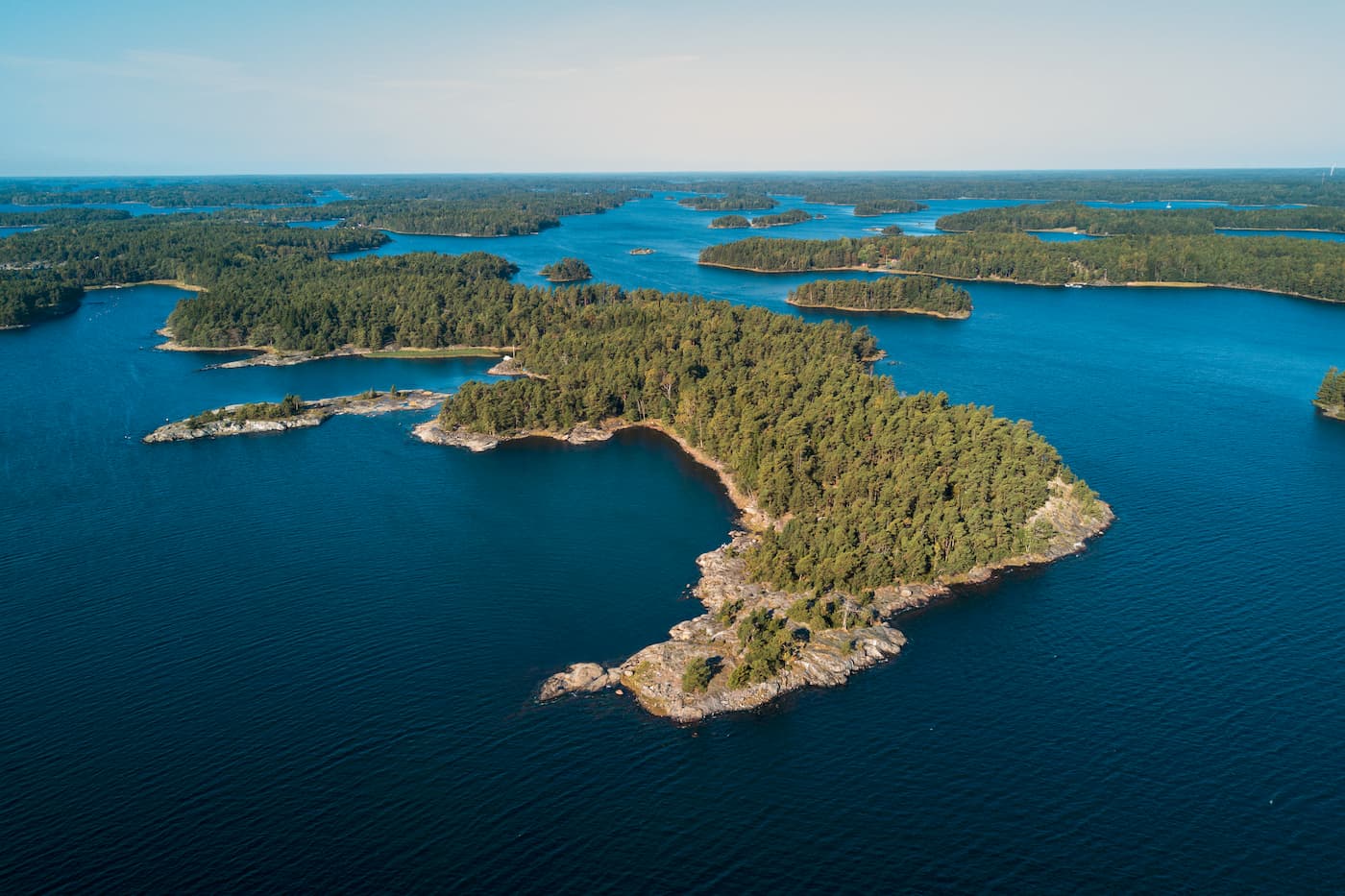 Flygbild över en skogbeklädd ö omgiven av djupblått vatten, med flera andra små öar synliga i fjärran under en klar himmel.