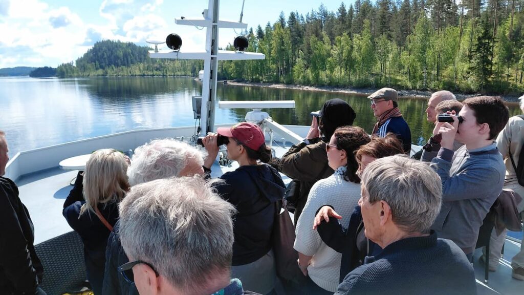 En grupp människor står på ett båtdäck och tittar ut över en lugn sjö och en skogsklädd strandlinje, några använder kikare under en delvis molnig himmel.