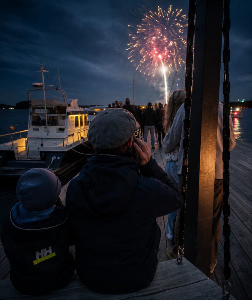 En grupp människor på en brygga tittar på ett fyrverkeri över vattnet på kvällen, med båtar förtöjda i närheten.