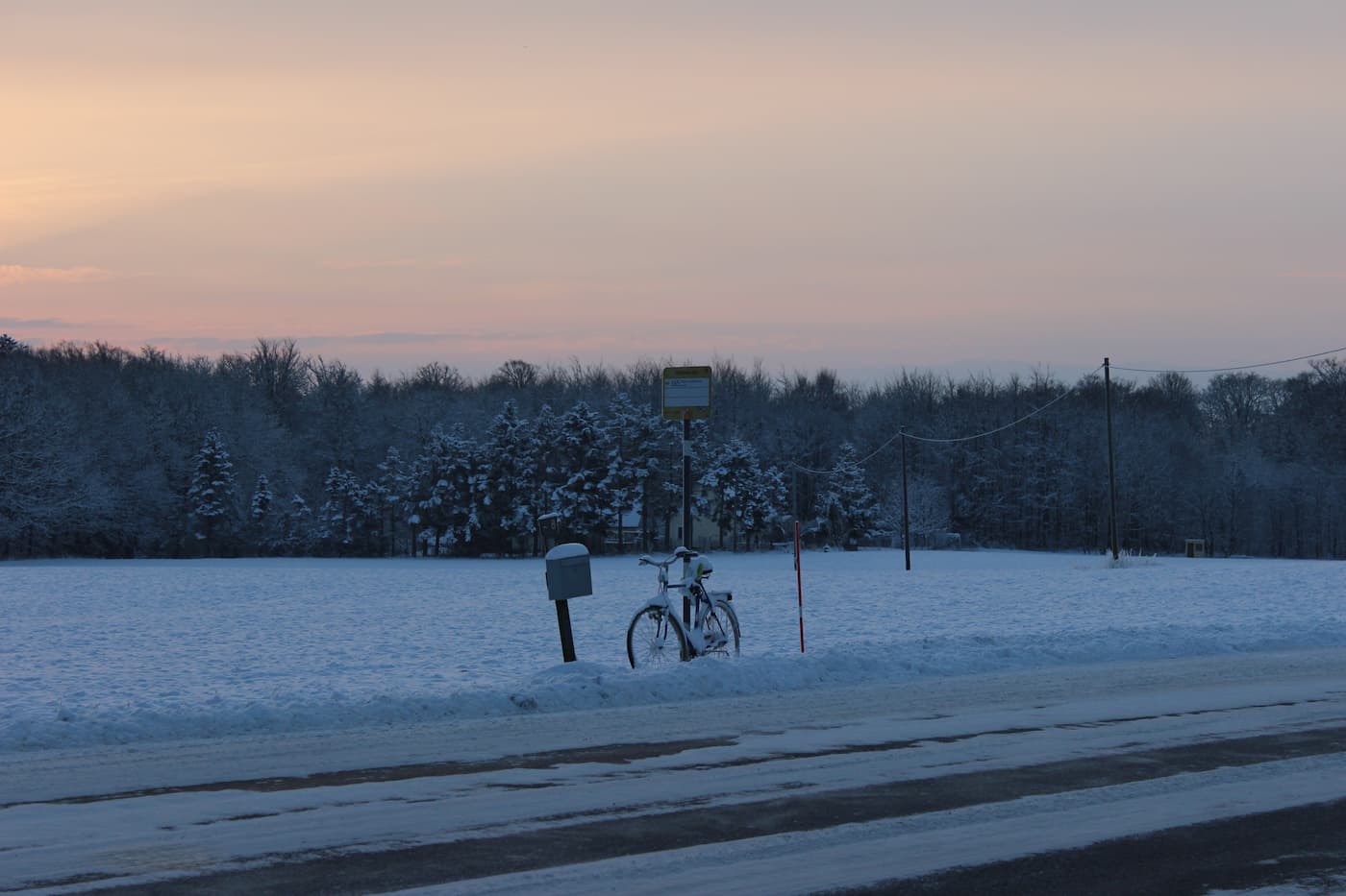En cykel står parkerad bredvid en brevlåda och en skylt vid en snötäckt vägkant med träd i bakgrunden vid solnedgången.