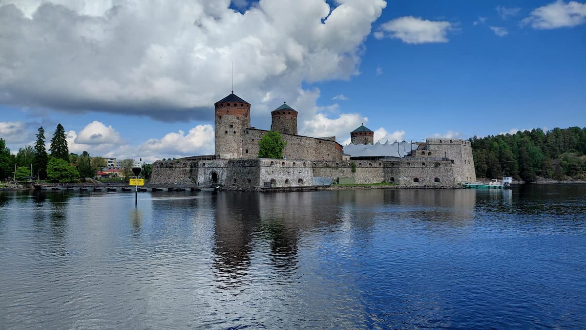 Ett stenslott med tre runda torn står vid vattnet, omgivet av träd och speglar sig i den lugna sjön under en delvis molnig himmel.