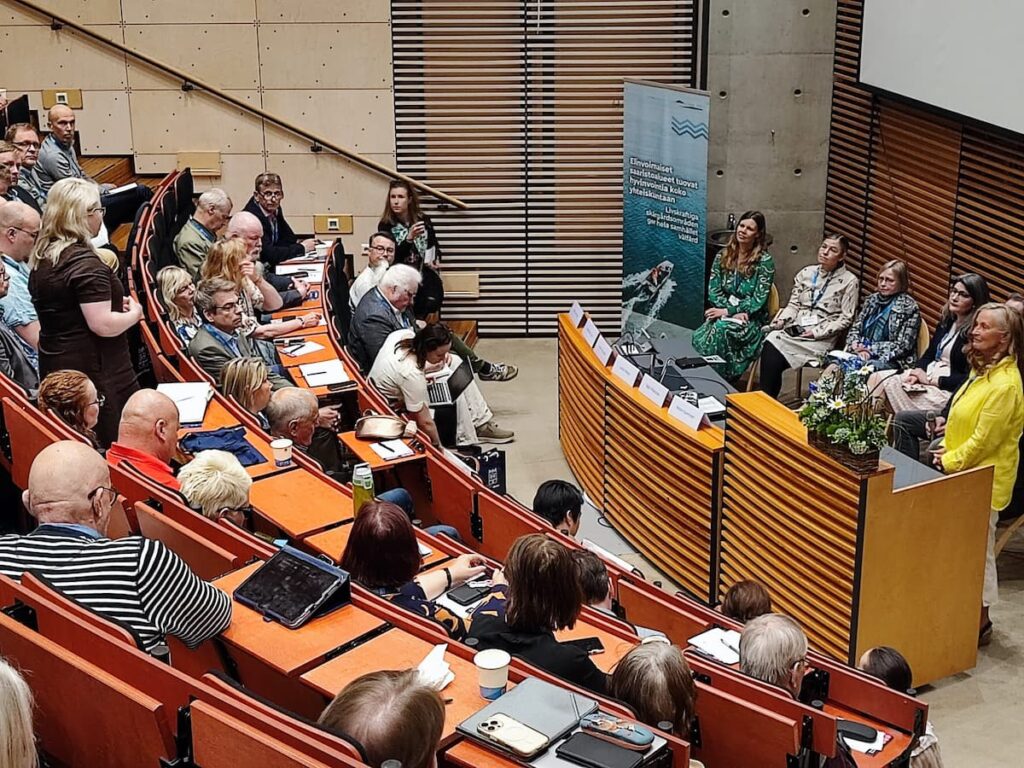 Personer som deltar i en paneldiskussion i en föreläsningssal; några sitter och antecknar, medan några står upp och talar. Paneldeltagarna sitter längst fram nära en presentationsskärm.